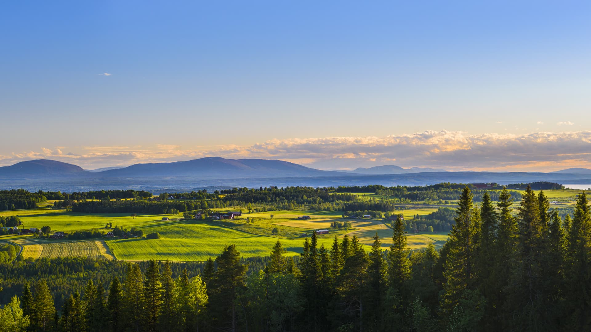 Solnedgång över ett jämtländskt landskap med skog och åkrar i förgrunden och fjäll och vattendrag i bakgrunden.